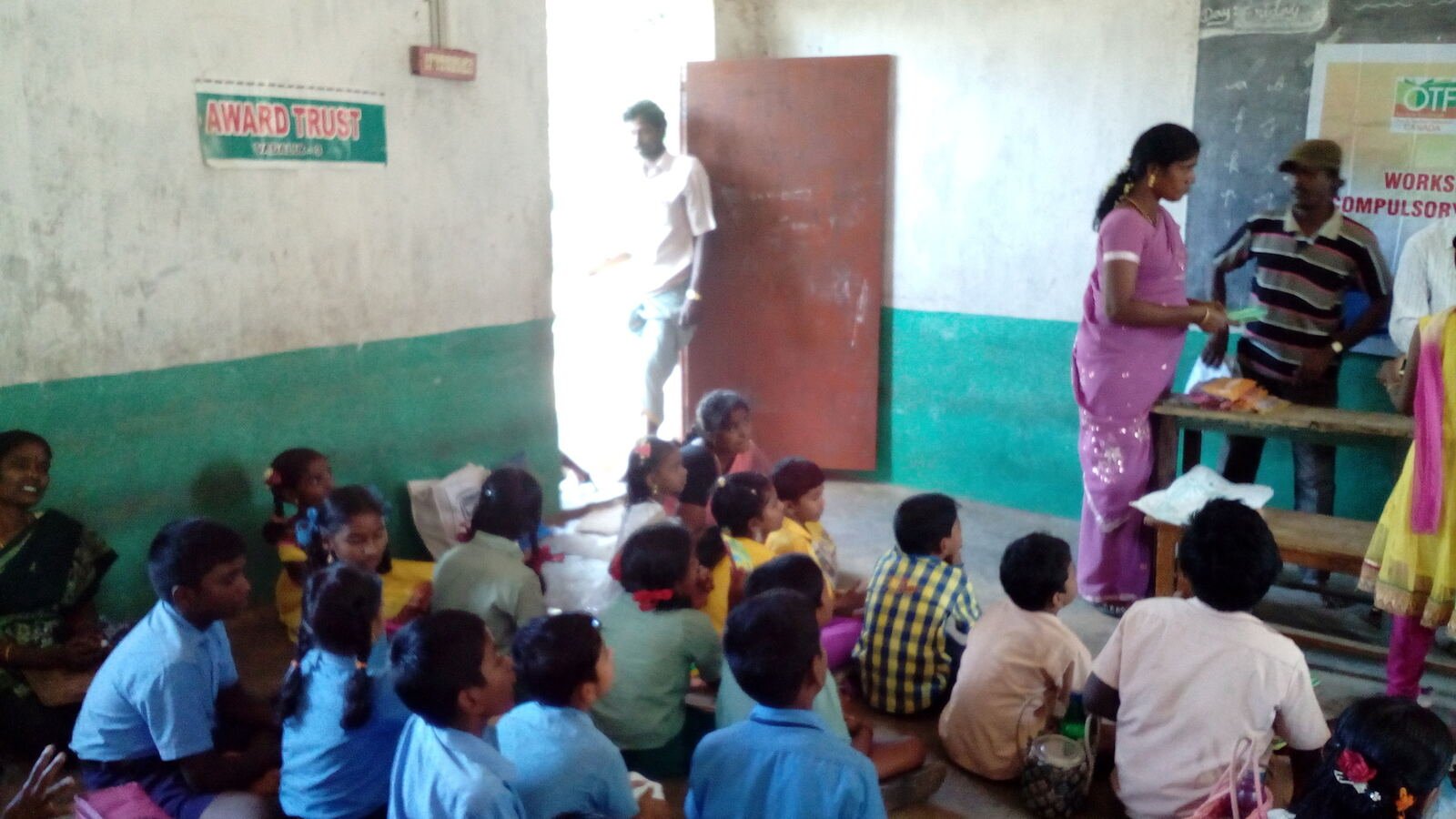 An AWARD TRUST signboard on a classroom wall, with children seated for a session